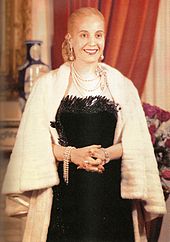 A picture of a Evita, former first lady of Argentina. Her hair is drawn into a tight bun at the back. She is wearing a black, low-cut dress. Around her neck là một number of chains. The lady's hands are folded in her front and she has a white fur shawl around her.