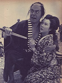 Jun Tazaki holding costar Michiko Saga and pointing a dagger at her while making a fierce face