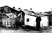 Hatted man stands outside shack