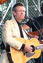 A man in a lght-coloured jacket playing a guitar and singing into a microphone