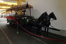 Two black horse statues stand harnessed to a 19th-century horse-drawn tram. The tram is open-air, with a canopy but no windows.