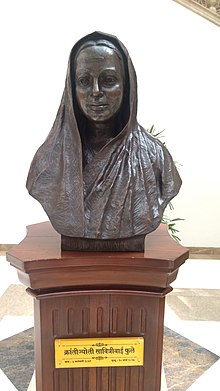 Bust of Savitribai Phule. The bust is in black color, the pedestal its on is made out of wood. The name plate on pedestal says "Kranjyoti Savitribai Phule" which roughly means "Revolutionary light Savitribai Phule" in Hindi. She is depicted to be wearing a saree.