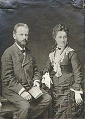 A middle aged man with dark hair and a beard, wearing a dark suit and holding a book, sits next to a young woman in a black dress wearing her hair up on her head