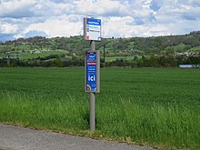 Photographie en couleurs du poteau d’un arrêt de bus au bord d'une route, environné uniquement de champs verts.