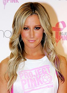A woman with long blonde hair wearing a white t-shirt with "PROJECT PINK" written on it while smiling to the camera.