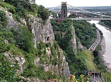 Rocky side to a gorge with a platform in front of a cave half way up. To the right are a road and river. In the distance are a suspension bridge and buildings.