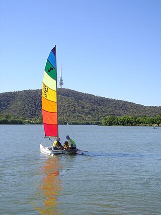 Catamaran with two adults and a child aboard. It has a rainbow-coloured horizontal striped sail. The water is calm and empty and Telstra Tower is in the background.