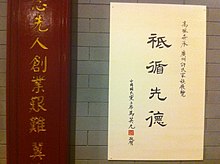 Deska na zdi s čínským nápisem ve sloupci ze čtyř znaků, po stranách malým písmem sloupce s doprovodným textem