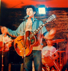 Zpívající muž s kytarou, v pozadí další dva hudebníci.