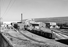 Black and white photo showing tracks and box cars waiting outside rows of rectangular shop buildings, one with a smoke stack.