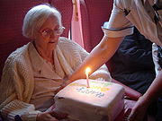 Seated elderly woman in glasses and sweater, viewing a birthday cake being placed in front of her by a nurse. The square cake has a single lit candle.