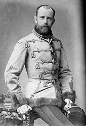 Photographie d'un homme barbu, semi-assis sur une table portant une veste militaire ornée de passants et bordée de fourrure, son regard est mélancolique et sa tête est assez dégarnie.