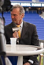 A grey-haired man in a black suit and yellow tie looks to his right, a microphone marked "MSG" held to his mouth