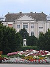 A colour photograph of Kálmán Tóth's memorial, front centre, before it several wreaths and flowers, surrounded by evergreen trees, and in the background a light stone building with Georgian escarpment