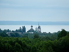 Photographie par temps couvert depuis les cimes d'une forêt d'églises et maisons d'un village bordant une grande étendue d'eau en arrière-plan.