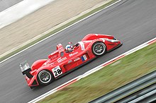 Photographie d'une voiture de sport-prototype rouge, noire et blanche, vue de haut et de profil, sur une piste.