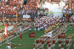 A large group of football players tightly huddled together wearing dark orange jerseys and white pants and helmets run onto the field surrounded on both sides by the marching band.