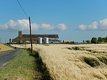 Au premier plan, des champs de céréales ; au second plan, un silo ; à gauche, une route étroite, qui est une ancienne voie romaine ; à droite on aperçoit la route D137.