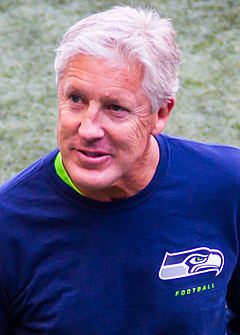 Цветная фотография головы и плеч седовласого Пита Кэрролла в темно-синей спортивной рубашке с логотипом Seattle Seahawks на левой груди.