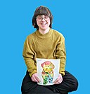 The image features a woman (Rita Winkler) sitting against a blue background, smiling broadly. She is wearing glasses with a rounded frame, a mustard yellow knit sweater, and black pants. The woman is holding a colorful painting of a whimsical character with a rounded head, decorated with patterns and stars. She has characteristics of Down syndrome such as almond-shaped eyes and a flat profile.