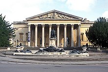  A Palladian style nineteenth century stone building with a large colonnaded porch. In front a large metal statue on a pedestal and fountains with decorations.