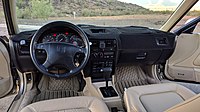 Carpeted dashboard of a left-hand-drive car with leather seats, an automatic gearbox, compact cassette player, heater and air conditioning with rotary controls, and an analogue gauge cluster.