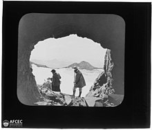 Photographie montrant deux hommes à l'entrée d'une grotte, prise depuis le fond de la grotte, le paysage figurant en arrière-plan.