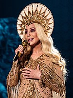 A woman dark eyes, holding a gold microphone stands on stage in a gold outfit
