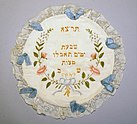 A white circle of fabric trimmed with lace, blue ribbons, and embroidery, including embroidered Hebrew letters