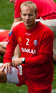A blond Caucasian man wearing red football kit with his right knee raised.