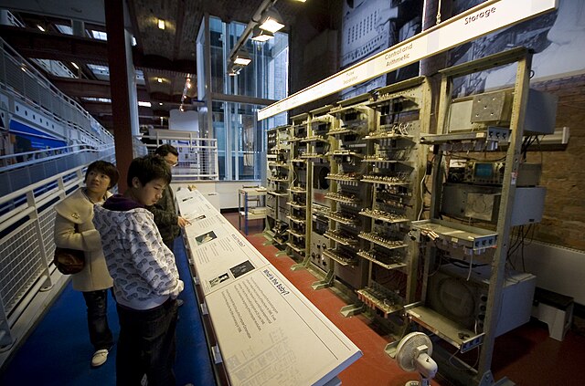 A series of seven tall metal racks filled with electronic equipment standing in front of a brick wall. Signs above each rack describe the functions carried out by the electronics they contain. Three visitors read from information stands to the left of the image.