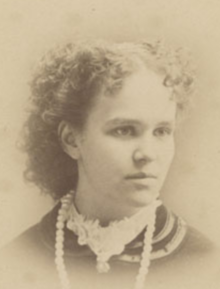 A young white woman with curly hair parted center. She is wearing a high frilly white collar and a strand of large, light-colored beads.