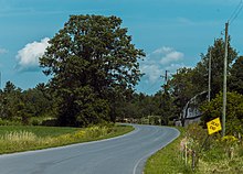 An unmarked paved two-lane road curves past a tree and barn, with a dead-end sign at right foreground, through countryside under clouds and blue sky during the summertime.