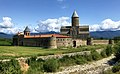 Depuis un chemin empierraillé, vue d'un grand monastère enceint d'une muraille en pierres de taille encerclant une église surmontée d'une tour cylindrique et coiffée d'un toit gris, le bâtiment est placé devant des montagnes bleuâtres sous un un ciel bleu où apparaissent quelques cumulus blancs.