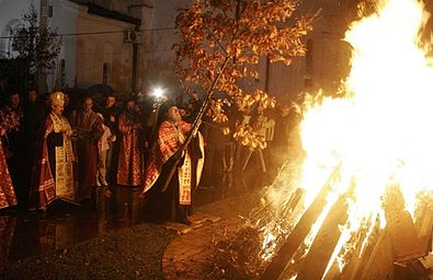 Open-air fire, built with conically arranged long pieces of wood, blazes in the night. Orthodox priest places a long oak sapling with brown leaves on the fire. The priest and the fire are surrounded by a ring of people watching them. In the background, walls of a great church are visible.