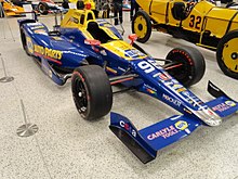 Victoire d'Indy Series, bleue et jaune, dans un musée, avec une voiture de course l'époque jaune à sa gauche.