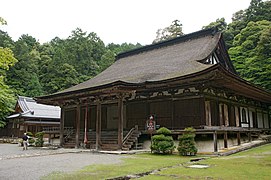 Photo couleur d'un bâtiment en bois à un étage d'un temple bouddhique doté d'un toit de chaume. À l'arrière-plan, une forêt d'arbres verts sous un ciel blanc laiteux.