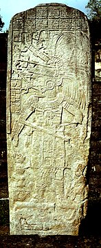 An upright stone shaft showing a figure looking to the left, holding a staff in one hand and wearing an elaborate feathered headdress. There are panels of hieroglyphs above and to the left of the figure.
