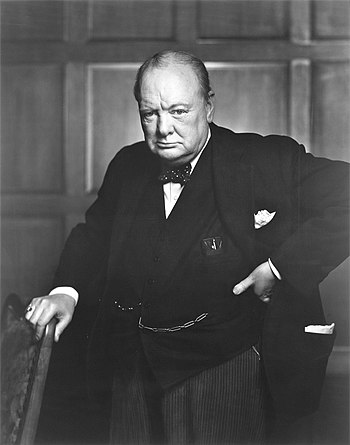 Black-and-white image of Winston Churchill standing with his left hand on his hip and his right hand resting on the back of a chair