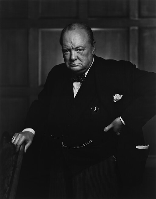 Churchill, aged 67, wearing a suit, standing & holding into the back of a chair