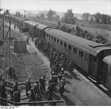 Uma foto em preto e branco de vários homens, alguns carregando mochilas, indo para um trem