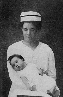 A woman in a nurse's uniform, looking down at and holding a baby with dark hair.