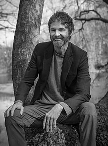 Black-and-white photo of George Dawes Green sitting outdoors by a tree, grinning at camera