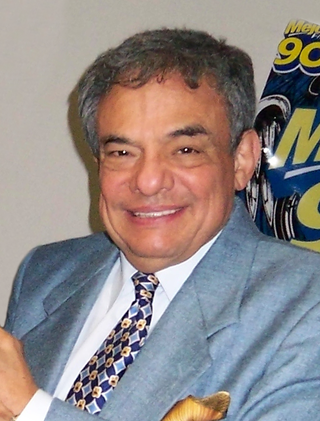 Man with grey hair wearing a light blue tuxedo is smiling at the camera.