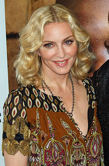 Upper body of a middle-aged blond woman. Her hair is parted in the middle and falls in waves to her shoulder. She is wearing a loose dress with black and brown prints on it. A locket is hung around her neck, coming up to her breasts. She is looking to the right and smiling.