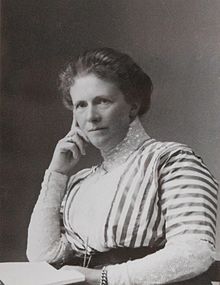 Black and white photograph of a woman. She is probably middle-aged. Her hair is in a fluffy bun on her head and she is wearing a striped gown, with a high polo-neck style lace collar. She is sat, leaning her head onto her right hand.