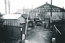 Une voiture ancienne s'apprête à emprunter un portail, derrière elle un bâtiment porte l'inscription « SHOCHIKU KINEMA KAMATA STUDIO » (image en noir et blanc).