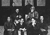 Posed group black-and-white photograph of seven Chinese men, possibly academics, in formal wear: two wear European-style suits, the five others wear Chinese traditional dress; four of the seven sit on the floor in the foreground; another sits on a chair behind them at center-left; two others stand in the background. They surround an eighth man who is robed, bearded, and sitting in a chair placed at center-left. Four elegant windows are behind them in a line.