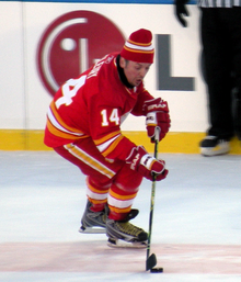 A hockey player in a red uniform with white and yellow trim. He is focused on the ice surface, as he attempts to control a puck while skating