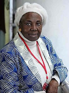 Trinidad Morgades Besari. Eine Afrikanische Frau in auffällig gemusterten afrikanischen Kleidern mit einer weißen Haube und Modeschmuck-Ketten.